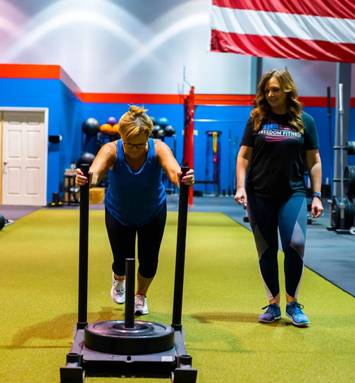 Woman pushing weighted sled with trainer support at Freedom Fitness gym